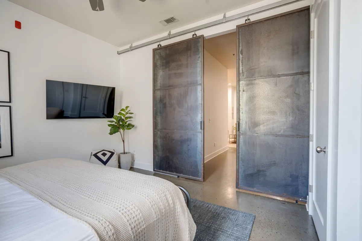 A calm, modern bedroom featuring a king-sized bed with textured bedding, a potted plant adding freshness, metal sliding doors for a contemporary touch, and a wall-mounted smart TV visible from the bed. Warm, natural light filters in, creating an inviting space for relaxation or unwinding with entertainment.