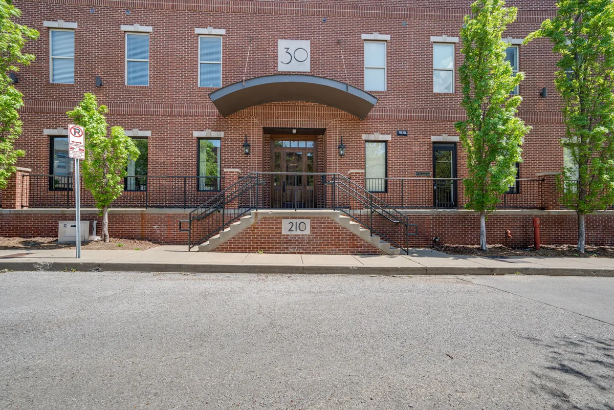 The image shows the stylish entrance steps of Urban Nest, large windows that let in ample natural light, and the distinctive brick facade with the number 30 clearly visible. The setting is inviting, with hints of communal activity around the entryway, suggesting a safe and welcoming arrival experience.