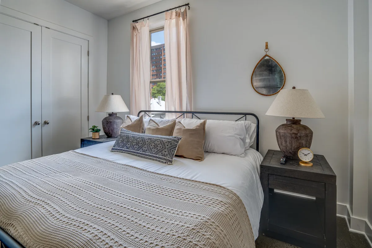 A cozy and serene bedroom showcasing a bed with decorative pillows, side tables topped with softly glowing lamps, and a window with sheer curtains that lets in gentle daylight. The overall space feels peaceful and inviting—ideal for rest and relaxation.