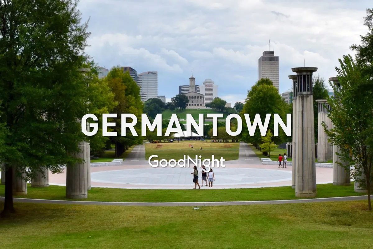 The image captures a central plaza bustling with people walking, set against the skyline of Germantown and framed by lush greenery and trees. Families can be seen enjoying casual strolls and picnics, reflecting a vibrant and welcoming community atmosphere.