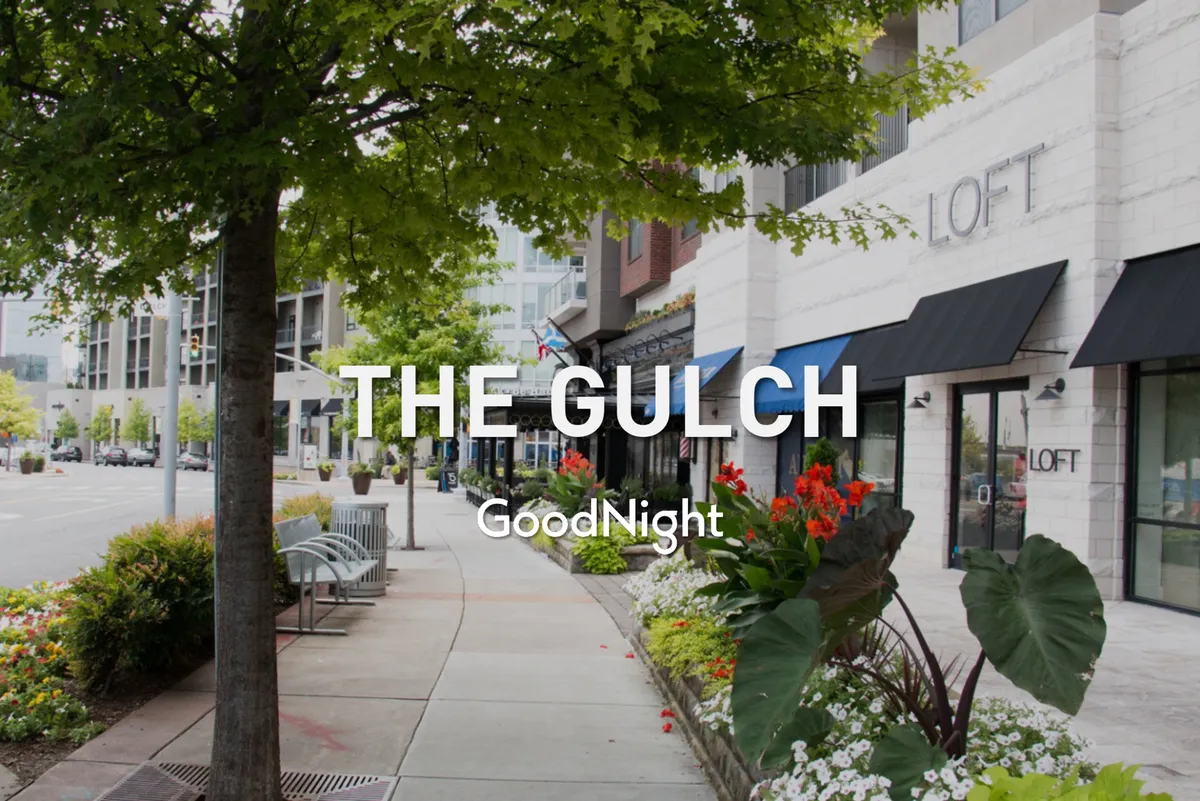 A charming street scene with benches, flower beds, trees, and inviting storefronts, capturing the essence of a walkable, lively Nashville neighborhood. The setting is ideal for evening strolls, stepping into intimate restaurants, or browsing local shops with a loved one.