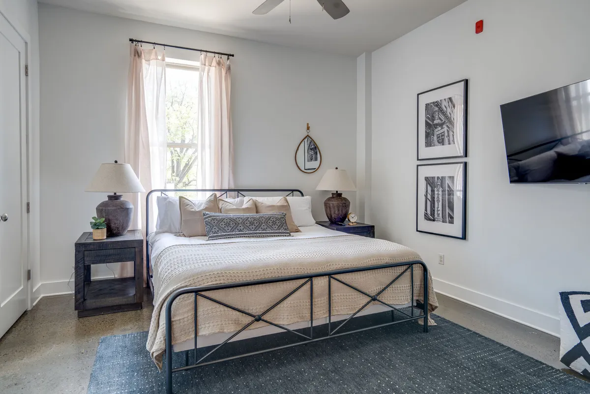The image showcases a stylish, sunlit bedroom with a plush bed adorned with decorative pillows, chic side tables, modern artwork above the headboard, and a large window framing natural light that fills the space with a tranquil glow. The decor exudes urban sophistication and comfort, inviting guests to relax and unwind.