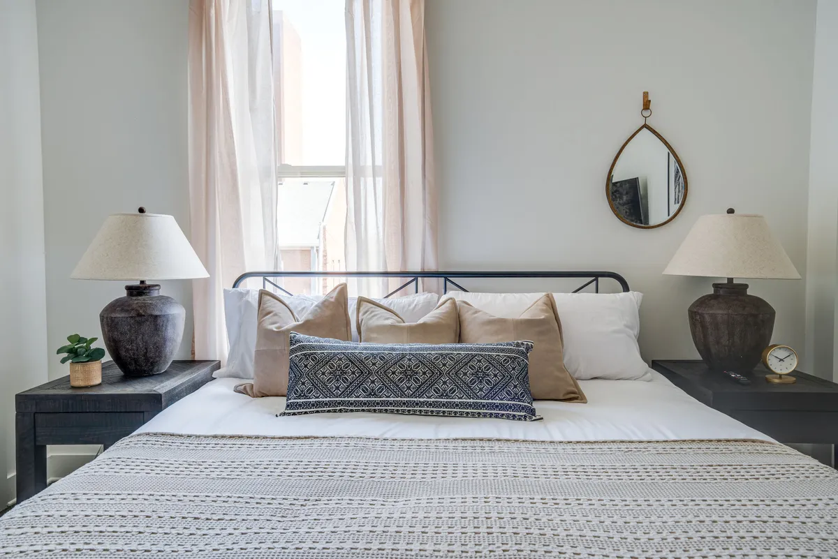 A serene bedroom with a plush bed dressed in decorative pillows, matching bedside lamps, and a wall-mounted mirror. The room is bathed in natural light, suggesting tranquility and privacy.