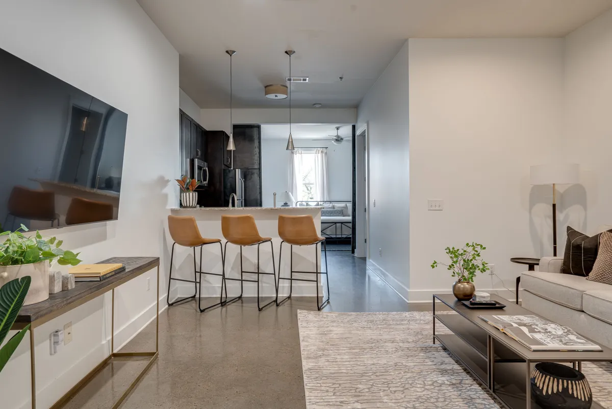 A modern open-plan living area with a plush sofa, bar stools at a sleek kitchen island, a large mounted smart TV, and a coffee table. The space is filled with natural light from expansive windows, and the decor is contemporary and inviting—ideal for both quiet relaxation and lively gatherings.